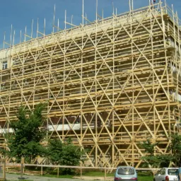 Budynek wielorodzinny w trakcie remontu elewacji, całkowicie otoczony drewnianymi rusztowaniami z widocznymi drabinkami, na pierwszym planie zieleń i zaparkowane samochody.