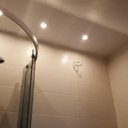 Wykończona łazienka z kabiną prysznicową z chromowanymi elementami, oświetleniem punktowym i beżowymi płytkami, widoczny wieszak na ścianie i fragment drzwi do innego pomieszczenia.