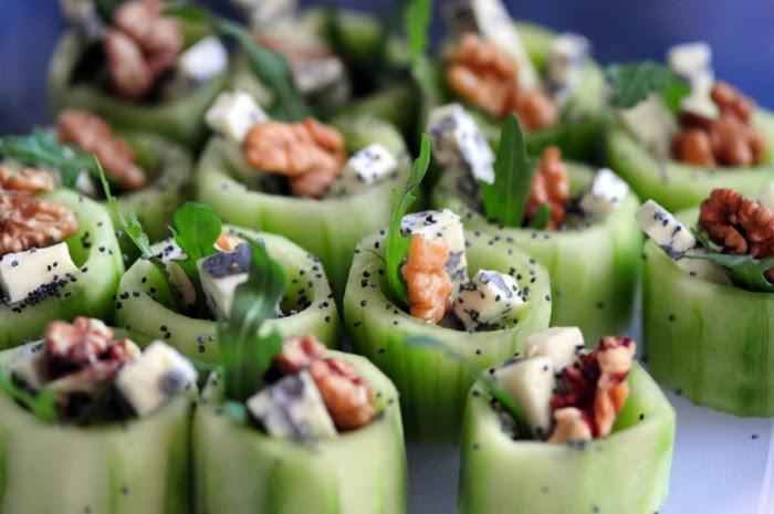 Dekoracyjne przekąski: rolki z ogórka z serem pleśniowym, orzechami włoskimi i rukolą, ułożone na niebieskim tle.