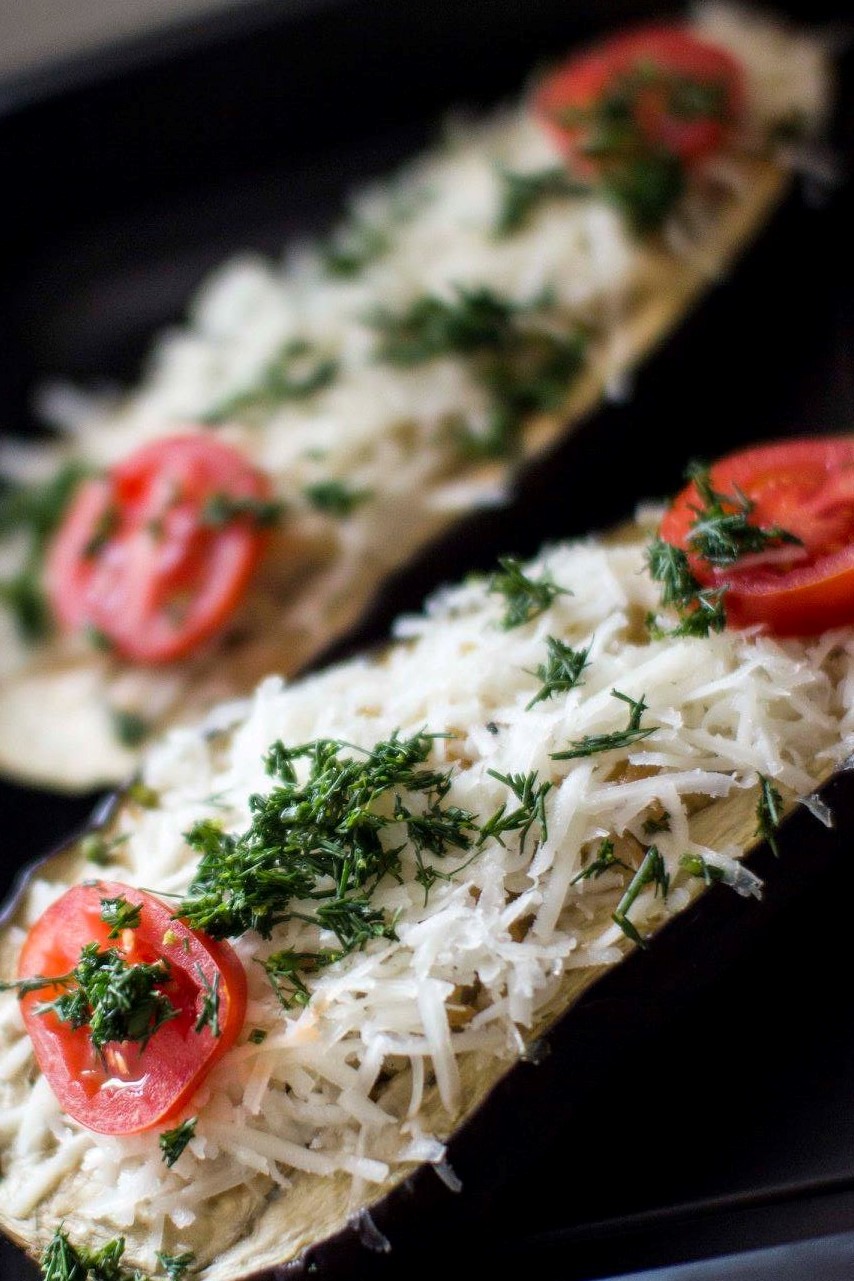 Dwa przekrojone bakłażany, wypełnione farszem, posypane startym serem i udekorowane plasterkami pomidora i świeżym koperkiem, ułożone na czarnej blasze.