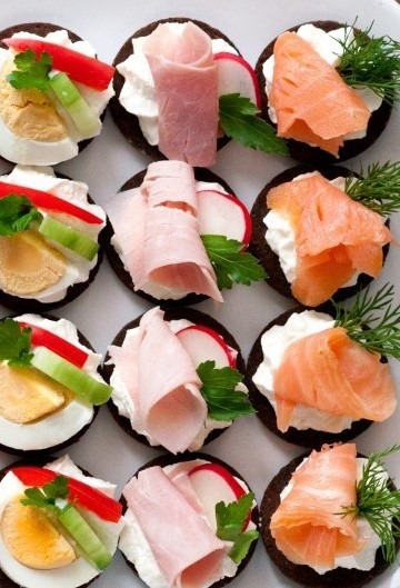 Koreczki na ciemnym pieczywie z kremowym serem, łososiem, szynką, jajkiem, rzodkiewką, ogórkiem i papryką, udekorowane koperkiem i natką pietruszki, ułożone na białym talerzu.