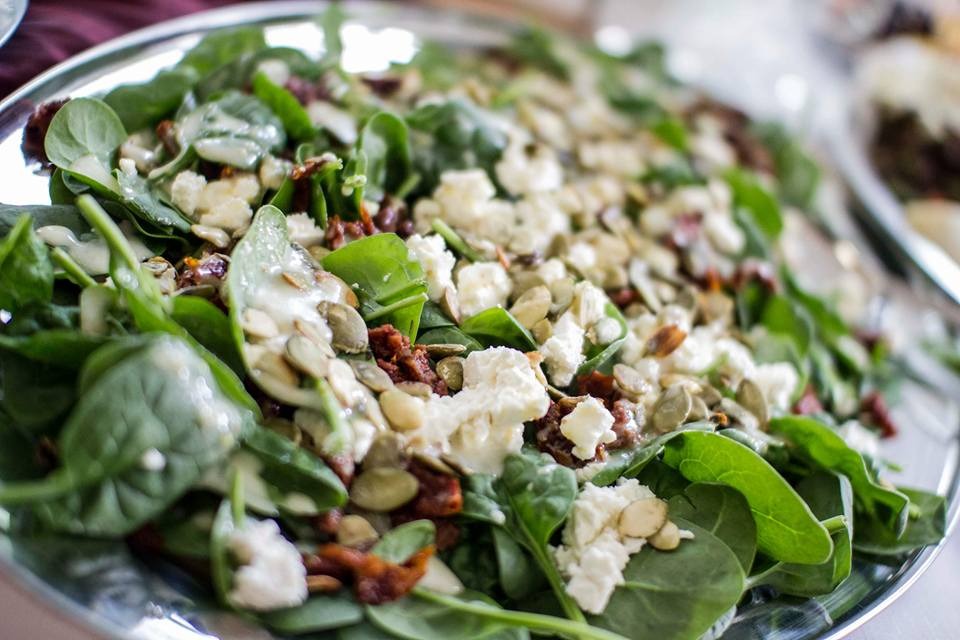 Sałatka ze szpinaku z suszonymi pomidorami, serem feta i pestkami dyni, polana sosem na srebrnej tacy.