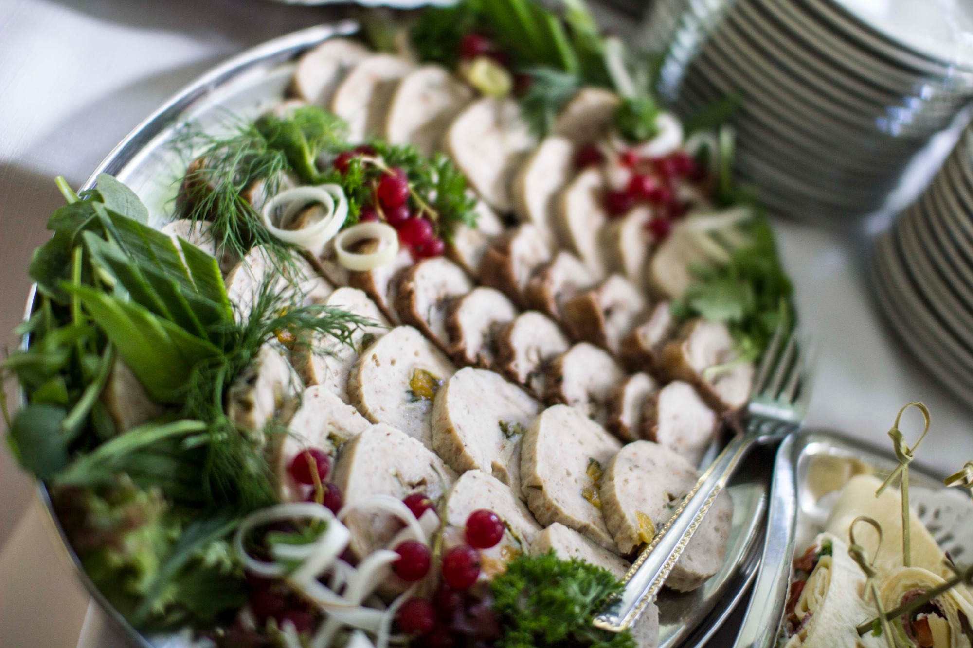 Srebrna taca z roladkami mięsnymi udekorowanymi zielonymi liśćmi, koperkiem, krążkami cebuli i czerwonymi porzeczkami, obok widoczne zwinięte tortille na małej tacce i stos talerzy.