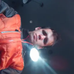 Portret mężczyzny w pomarańczowej kurtce z niskiej perspektywy, oświetlony od dołu, z widocznymi elementami sufitu w tle. Nietypowy kąt ujęcia.