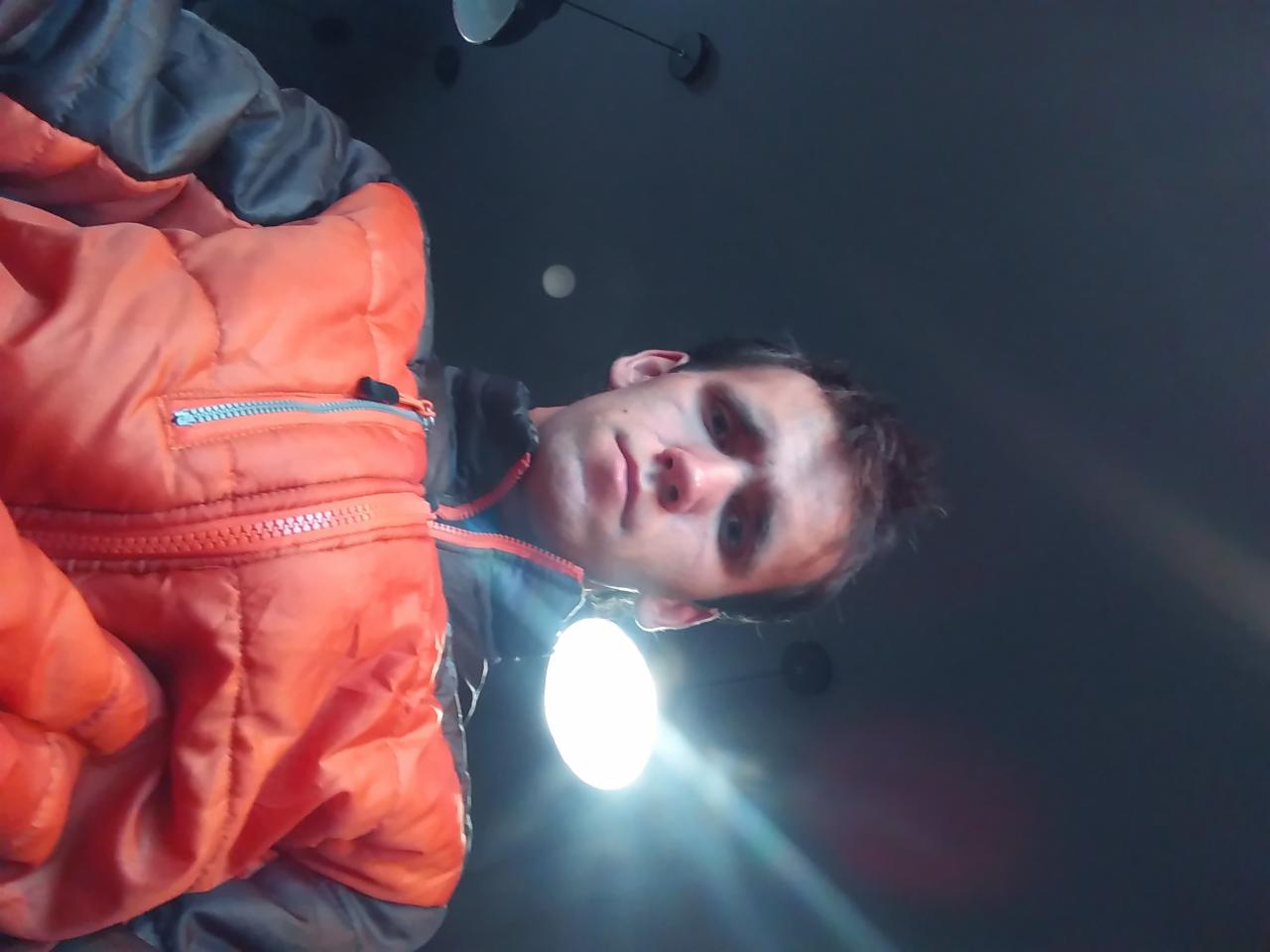 Portret mężczyzny w pomarańczowej kurtce z niskiej perspektywy, oświetlony od dołu, z widocznymi elementami sufitu w tle. Nietypowy kąt ujęcia.