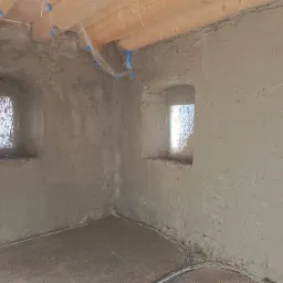 Surowe wnętrze z oknami w grubych murach, ściany pokryte świeżym tynkiem, zabezpieczone folią drewniane belki stropowe, widoczne przewody w podłodze.