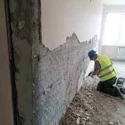 RAF-BUD Rafał Lisek - Pracownik w żółtej kamizelce i niebieskim kasku skuwa stary tynk ze ściany w pomieszczeniu w trakcie remontu, widoczne pozostałości tynku na podłodze, surowa ściana w tle.