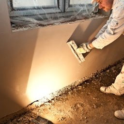 RAF-BUD Rafał Lisek - Pracownik w białym kombinezonie wyrównuje świeżo nałożony tynk na ścianie, widoczne okno zabezpieczone folią i zabrudzona podłoga.