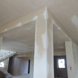 RAF-BUD Rafał Lisek - Wnętrze w trakcie prac wykończeniowych: surowe ściany z widocznymi niedoskonałościami, przygotowane pod malowanie, z widocznymi schodami w tle i oknem w oddali.