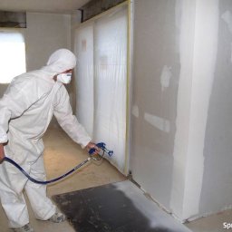 RAF-BUD Rafał Lisek - Pracownik w kombinezonie ochronnym nanosi tynk maszynowy na ścianę za pomocą pistoletu natryskowego, widoczne zabezpieczenia folią i niedoskonałości podłoża.