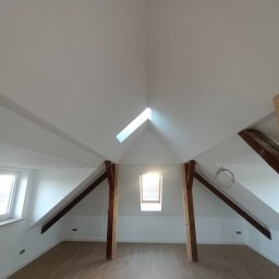 RAF-BUD Rafał Lisek - Poddasze po remoncie z drewnianymi belkami, nowymi oknami dachowymi i świeżo pomalowanymi ścianami na biało, widoczne gniazdka elektryczne i wystające kable.