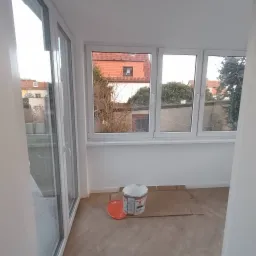 Pomalowane na biało wnętrze balkonu z widokiem na sąsiednie budynki, z widocznym wiadrem z farbą Alpinawell i kartonem zabezpieczającym podłogę.