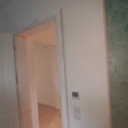 RAF-BUD Rafał Lisek - Świeżo pomalowane, białe wnętrze z zabezpieczonymi folią i taśmą malarską ościeżnicami drzwiowymi, widoczne gniazdko elektryczne i panel sterowania, fragment ściany wyłożony zieloną mozaiką...