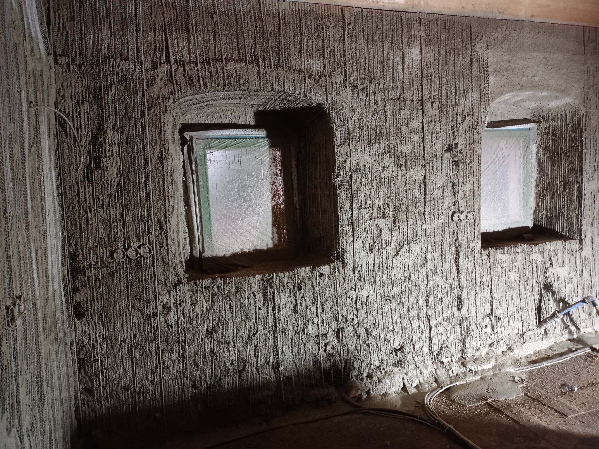 Ściana w trakcie nakładania tynku maszynowego z widocznymi dwoma oknami zabezpieczonymi folią, pionowymi rowkami w tynku i wystającymi przewodami elektrycznymi.