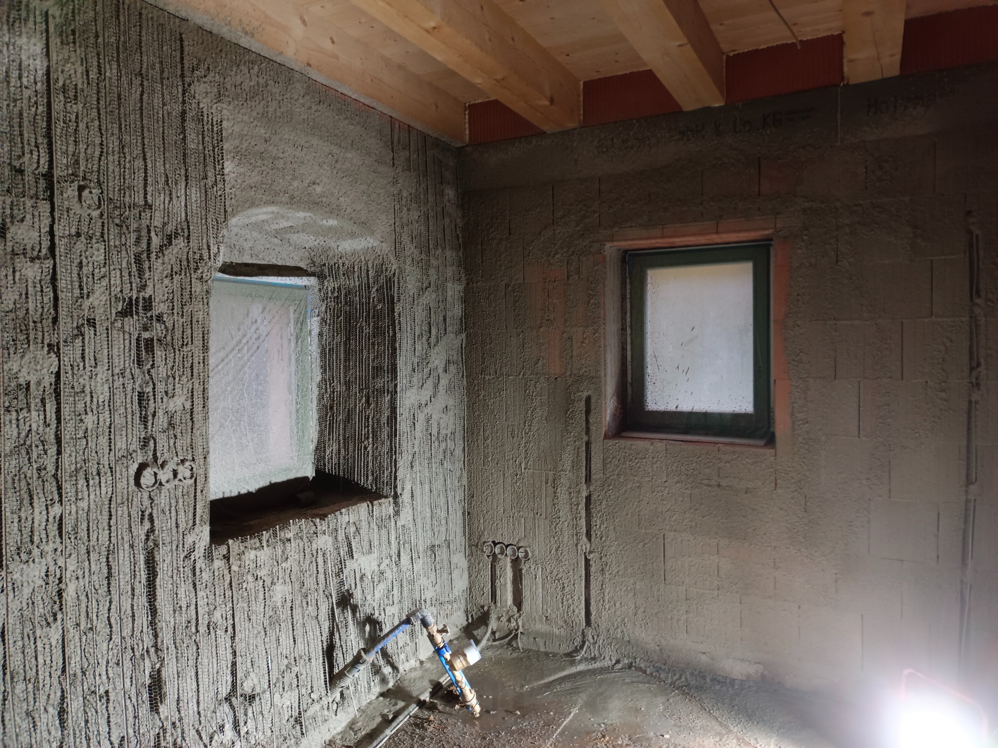 Wnętrze w trakcie prac tynkarskich: ściany pokryte świeżym tynkiem z widoczną siatką zbrojeniową, drewniany strop, okno z ciemną ramą, instalacja wodna przy podłodze, niedokończone wykończenie...