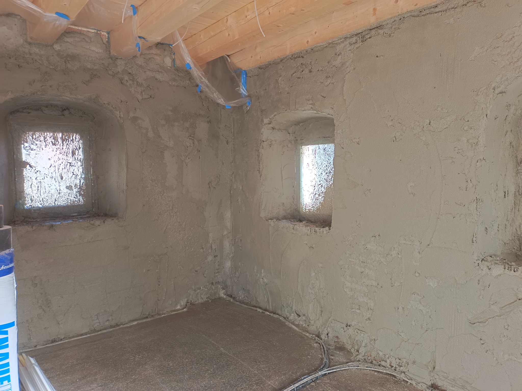 Surowe wnętrze z oknami w grubych murach, ściany pokryte świeżym tynkiem, zabezpieczone folią drewniane belki stropowe, widoczne przewody w podłodze.