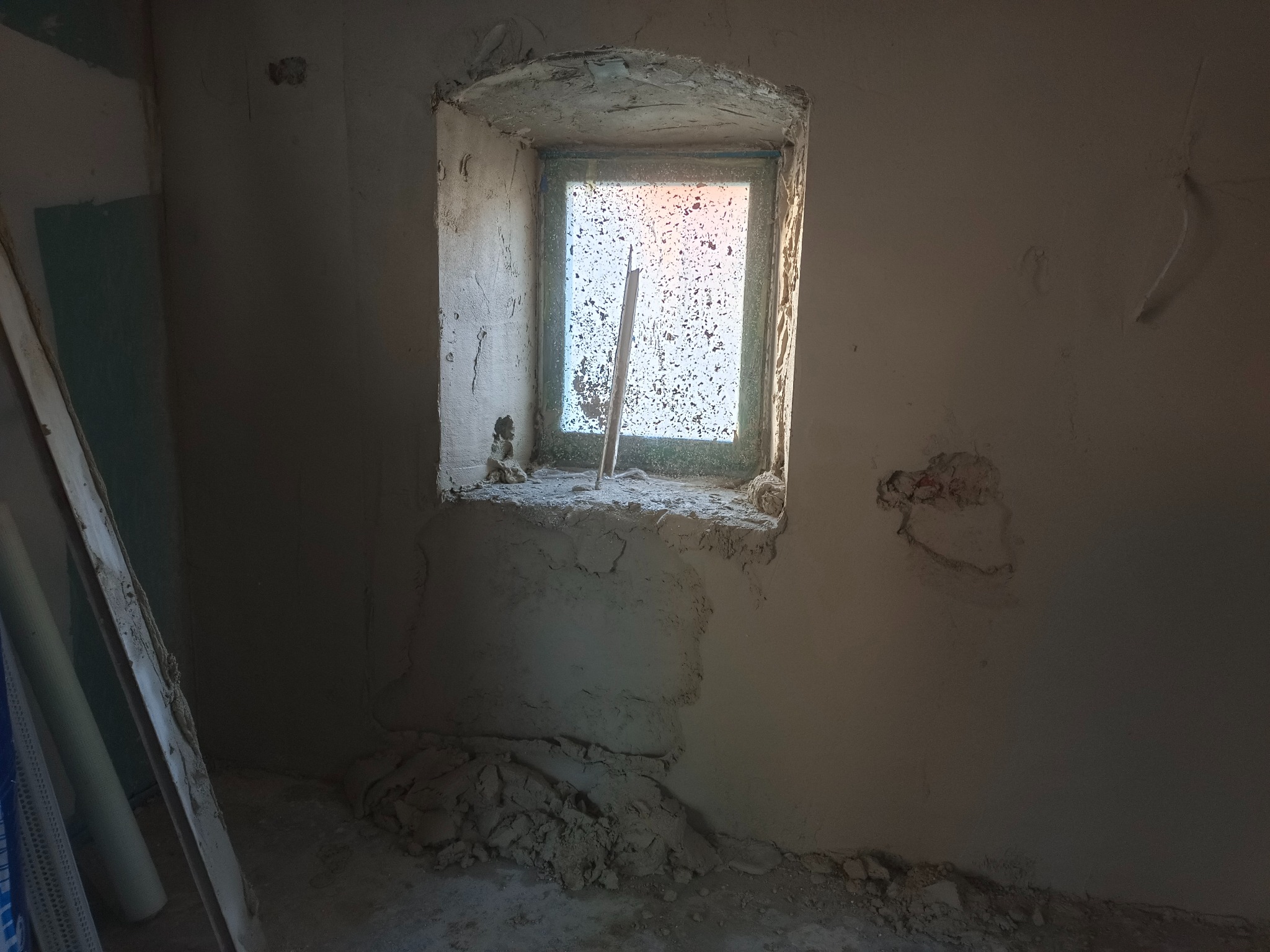Wnętrze w trakcie tynkowania maszynowego, widoczne surowe ściany, okno z zarysami tynku i resztki materiałów budowlanych na podłodze.