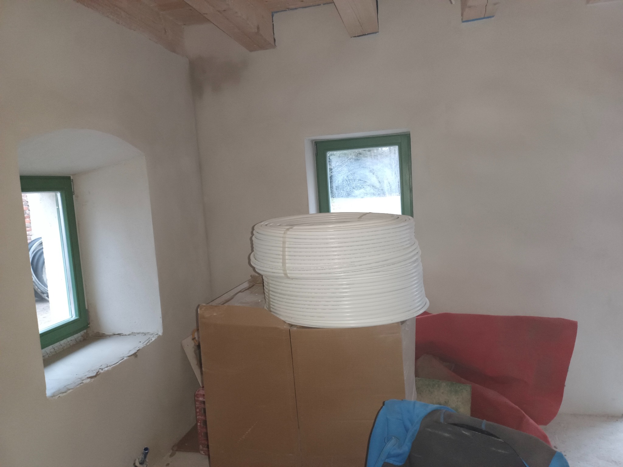 Wnętrze pokoju po tynkowaniu maszynowym; widoczne surowe ściany, drewniane belki stropowe, okna z zielonymi ramami oraz zwój białych rur na kartonowych pudłach.