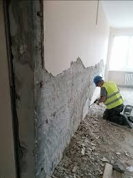 Pracownik w żółtej kamizelce i niebieskim kasku skuwa stary tynk ze ściany w pomieszczeniu w trakcie remontu, widoczne pozostałości tynku na podłodze, surowa ściana w tle.