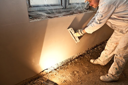 Pracownik w białym kombinezonie wyrównuje świeżo nałożony tynk na ścianie, widoczne okno zabezpieczone folią i zabrudzona podłoga.