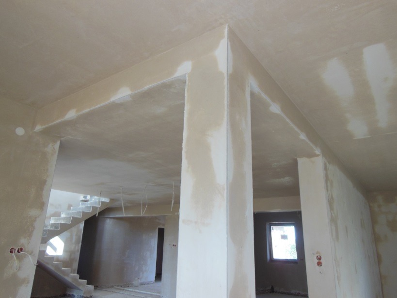 Wnętrze w trakcie prac wykończeniowych: surowe ściany z widocznymi niedoskonałościami, przygotowane pod malowanie, z widocznymi schodami w tle i oknem w oddali.