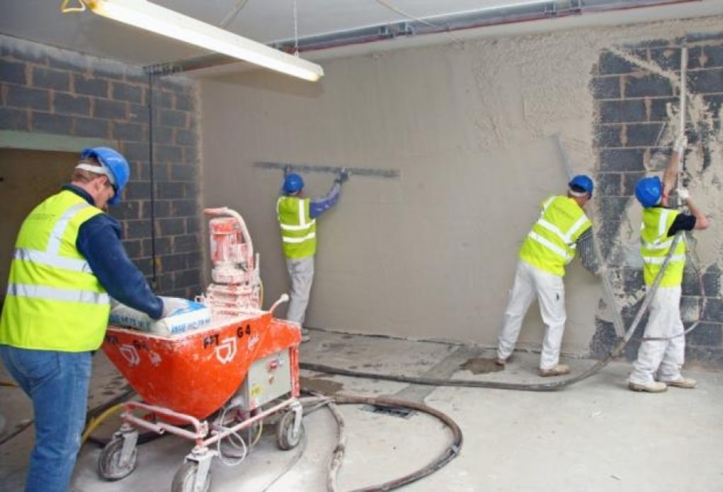 Brygada w kamizelkach odblaskowych nakłada tynk maszynowy na ścianę z cegły, używając pomarańczowej maszyny do tynkowania, widoczny wąż i worek z zaprawą.