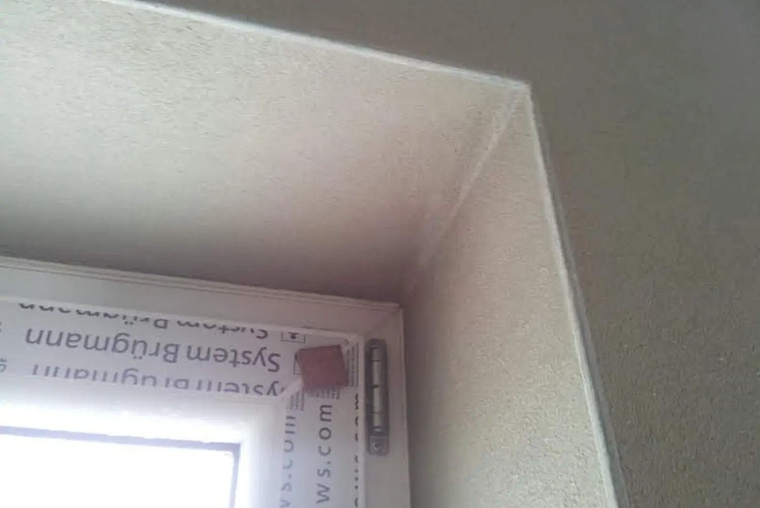 Narożnik okna po tynkowaniu maszynowym, widoczna struktura tynku i zabezpieczona rama okienna.