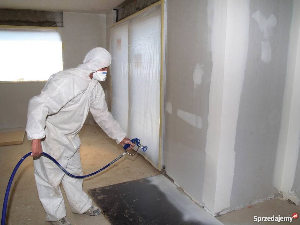 Pracownik w kombinezonie ochronnym nanosi tynk maszynowy na ścianę za pomocą pistoletu natryskowego, widoczne zabezpieczenia folią i niedoskonałości podłoża.