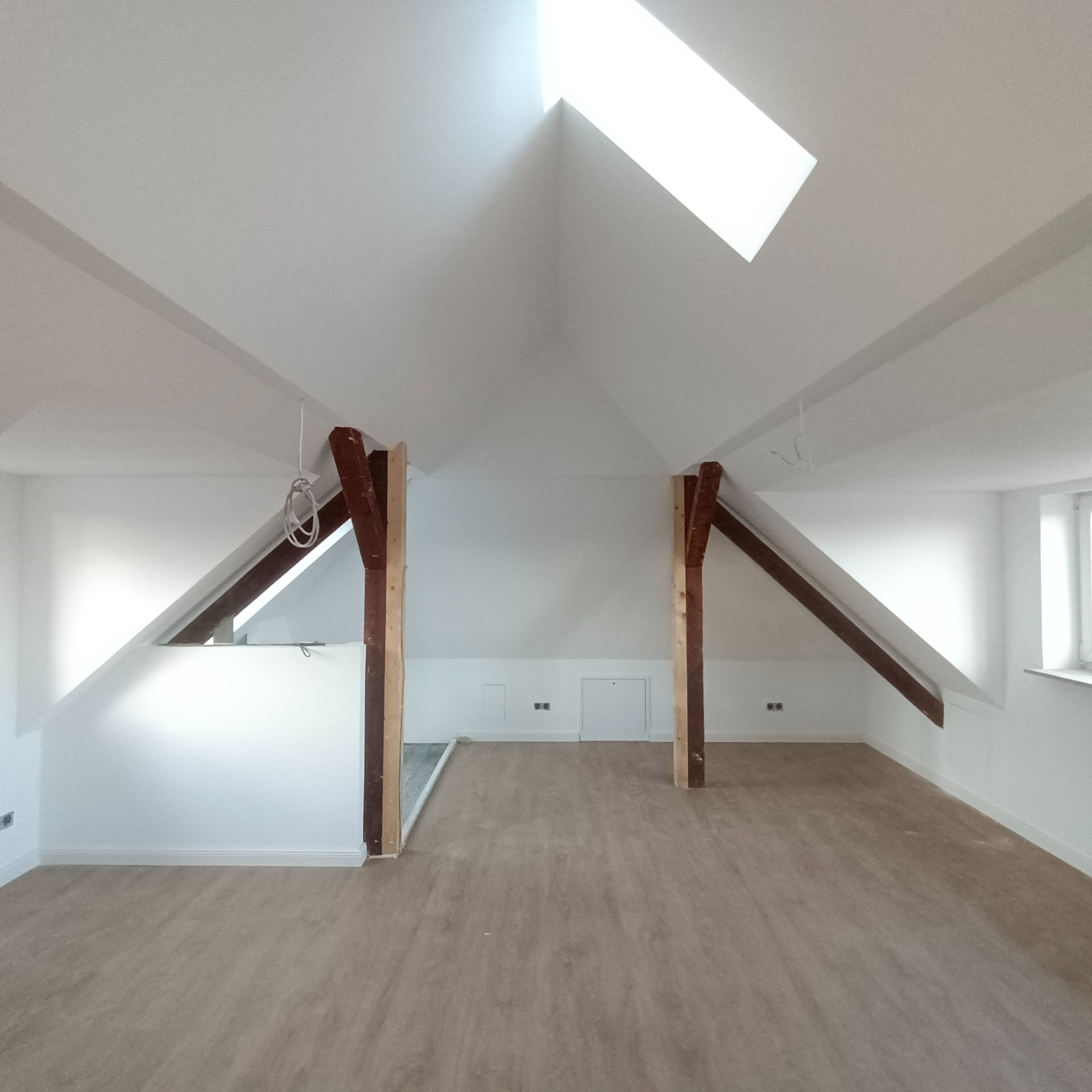 Wnętrze poddasza po remoncie z drewnianymi belkami, nową podłogą i oknem dachowym, świeżo pomalowane na biało.