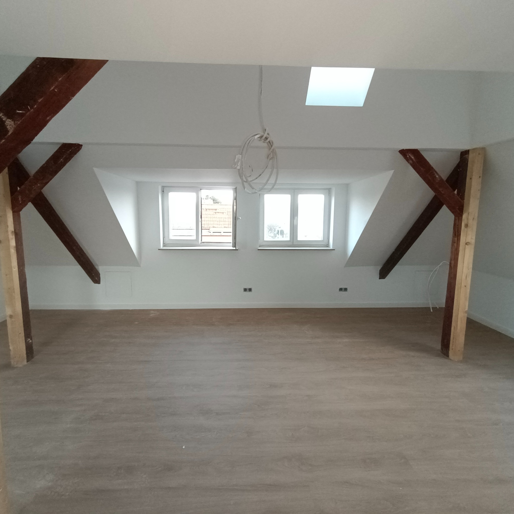 Poddasze z nową podłogą, surowe drewniane belki konstrukcyjne, dwa okna i świetlik w suficie, przewody zwisające z sufitu czekające na podłączenie.