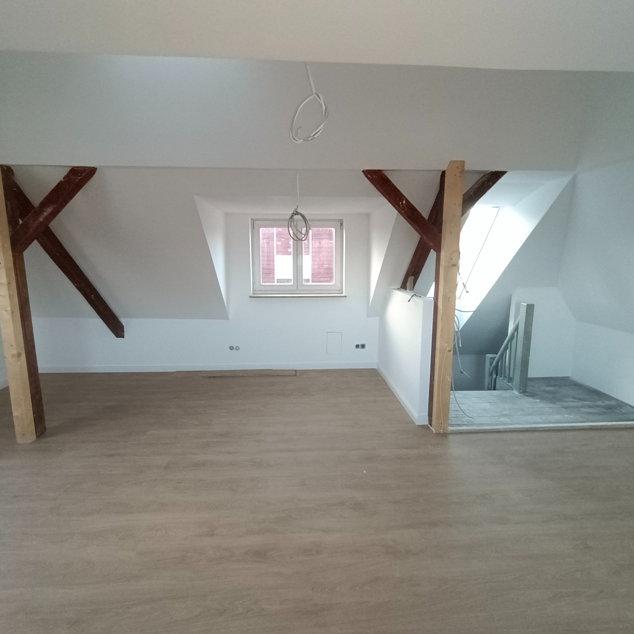 Poddasze z drewnianymi belkami, świeżo położoną podłogą, białymi ścianami i widocznymi przewodami elektrycznymi. W tle okno i schody.
