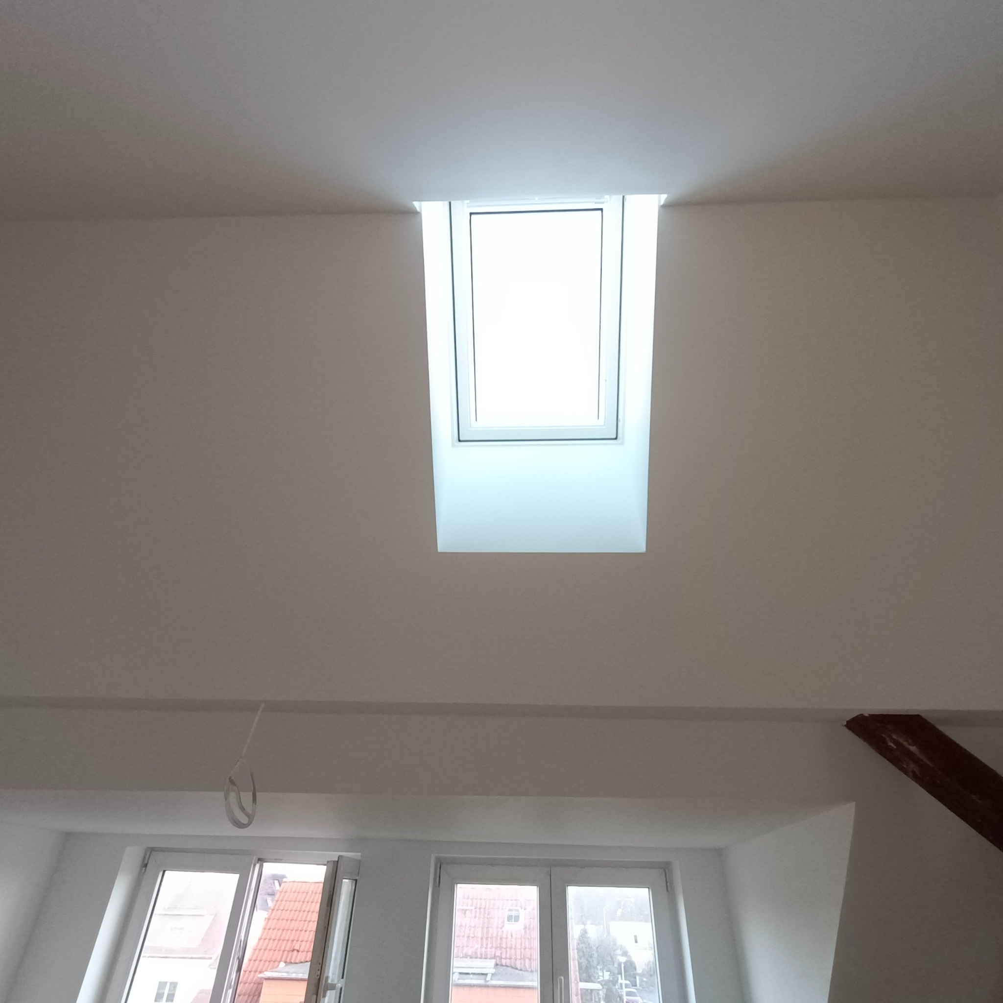 Wnętrze poddasza z oknem dachowym po remoncie, widoczne gładkie, białe ściany i sufit oraz fragment drewnianej belki stropowej.