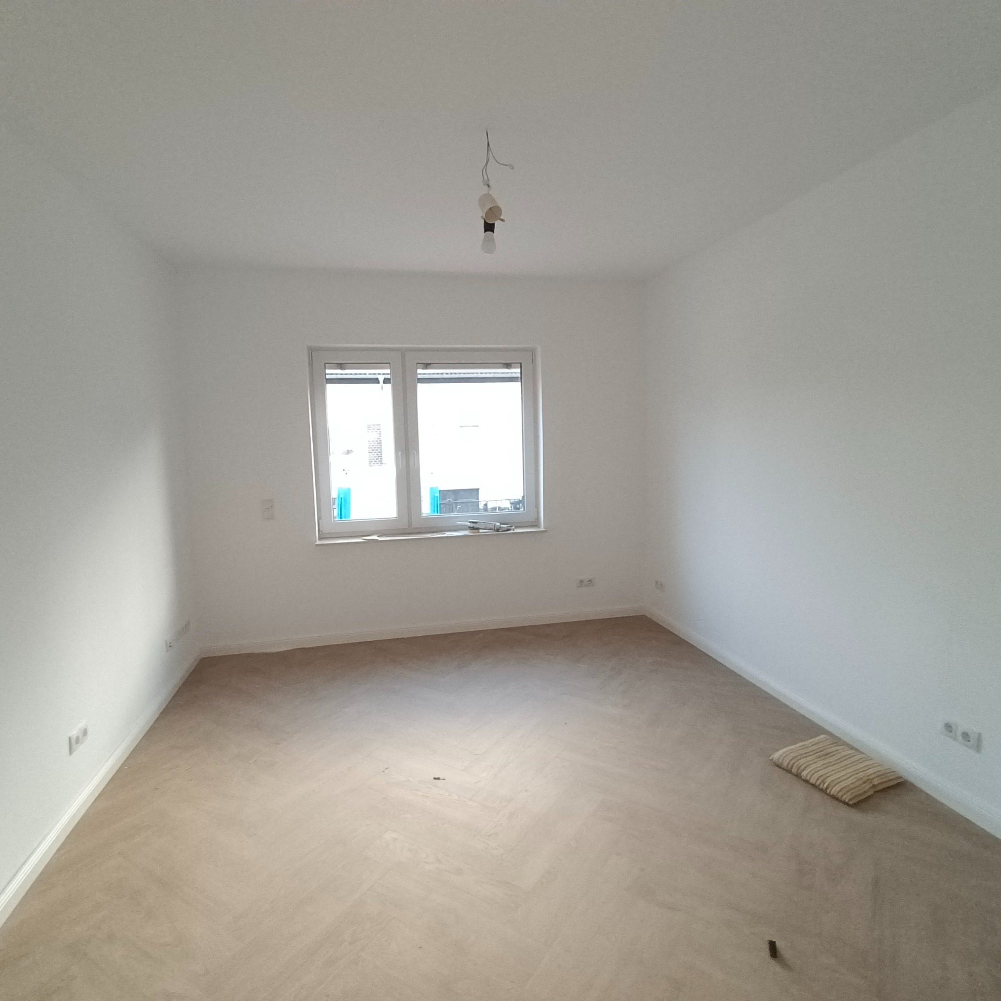 Pusty pokój po remoncie z jasnymi ścianami, parkietem w jodełkę i oknem z widokiem na ulicę, na suficie zwisający kabel z żarówką.