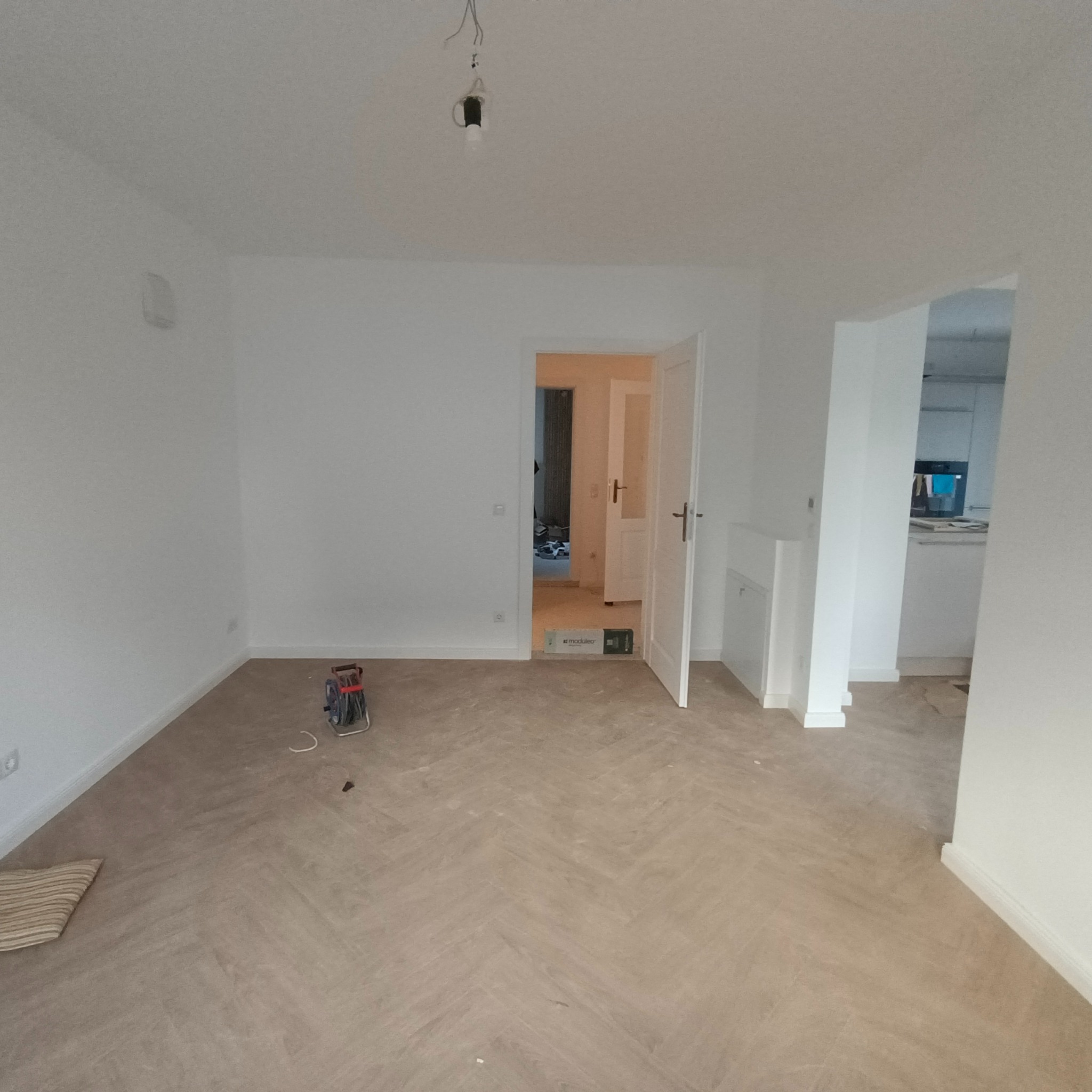 Wnętrze pokoju po remoncie z nową podłogą ułożoną w jodełkę, świeżo pomalowanymi ścianami na biało, widoczną puszką po żarówce zwisającą z sufitu i rozłożonym przedłużaczem na podłodze, z widokiem...