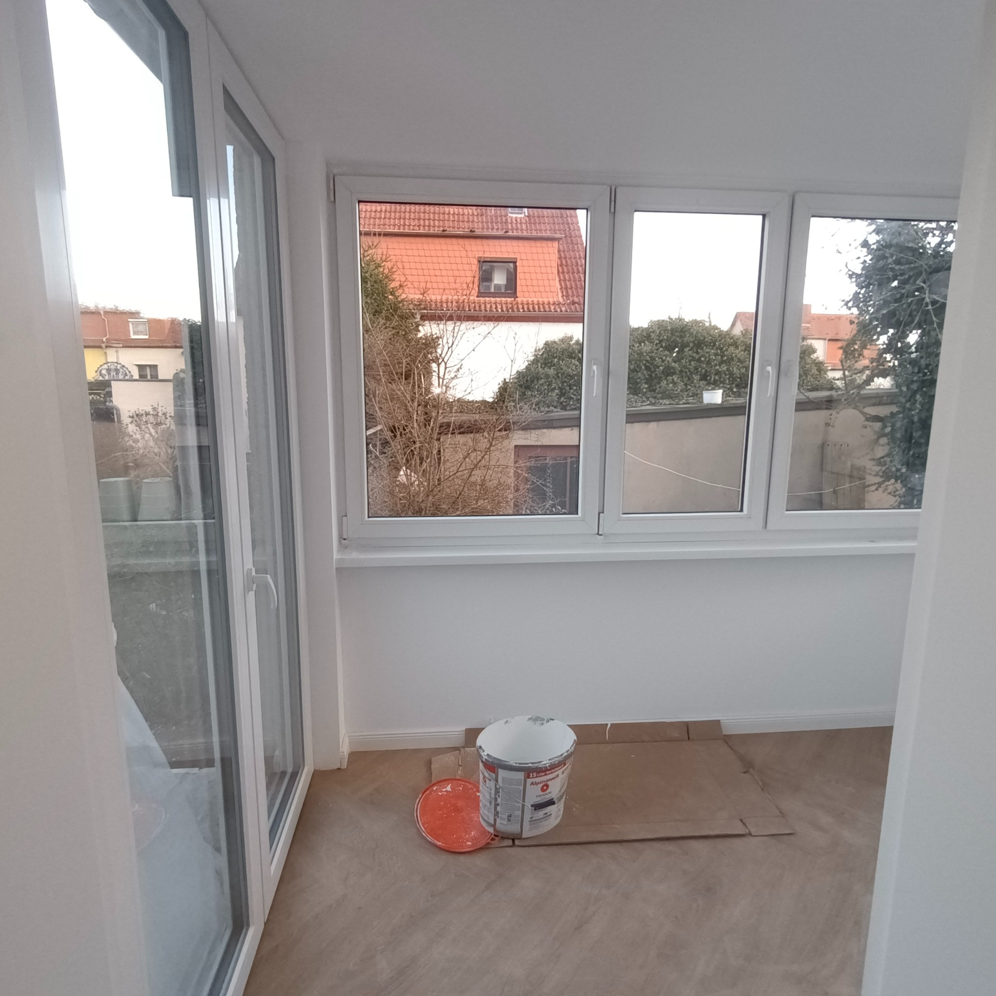 Pomalowane na biało wnętrze balkonu z widokiem na sąsiednie budynki, z widocznym wiadrem z farbą Alpinawell i kartonem zabezpieczającym podłogę.