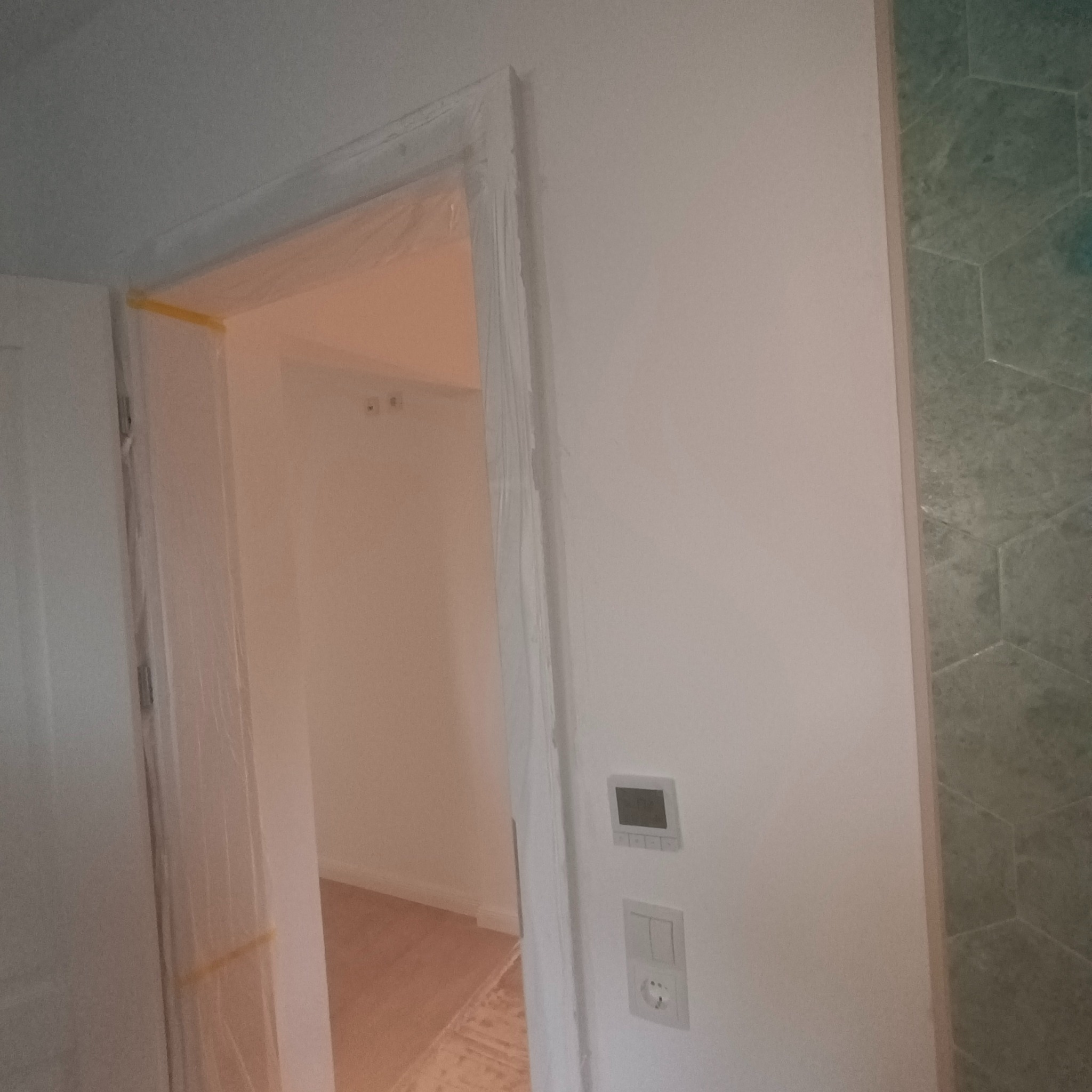 Świeżo pomalowane, białe wnętrze z zabezpieczonymi folią i taśmą malarską ościeżnicami drzwiowymi, widoczne gniazdko elektryczne i panel sterowania, fragment ściany wyłożony zieloną mozaiką...