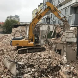 Żółta koparka VMG na gruzowisku podczas rozbiórki budynku w miejskiej scenerii, z widocznymi fragmentami ścian i cegieł.