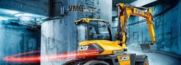 Żółta koparka JCB na tle betonowej konstrukcji z widoczną instalacją elektryczną i czerwonymi smugami światła, sugerująca prezentację możliwości maszyny.
