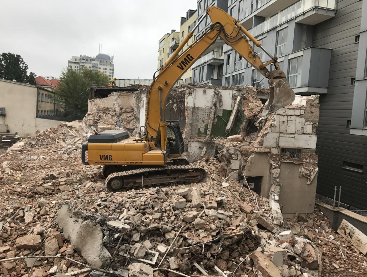 Żółta koparka VMG na gruzowisku podczas rozbiórki budynku w miejskiej scenerii, z widocznymi fragmentami ścian i cegieł.