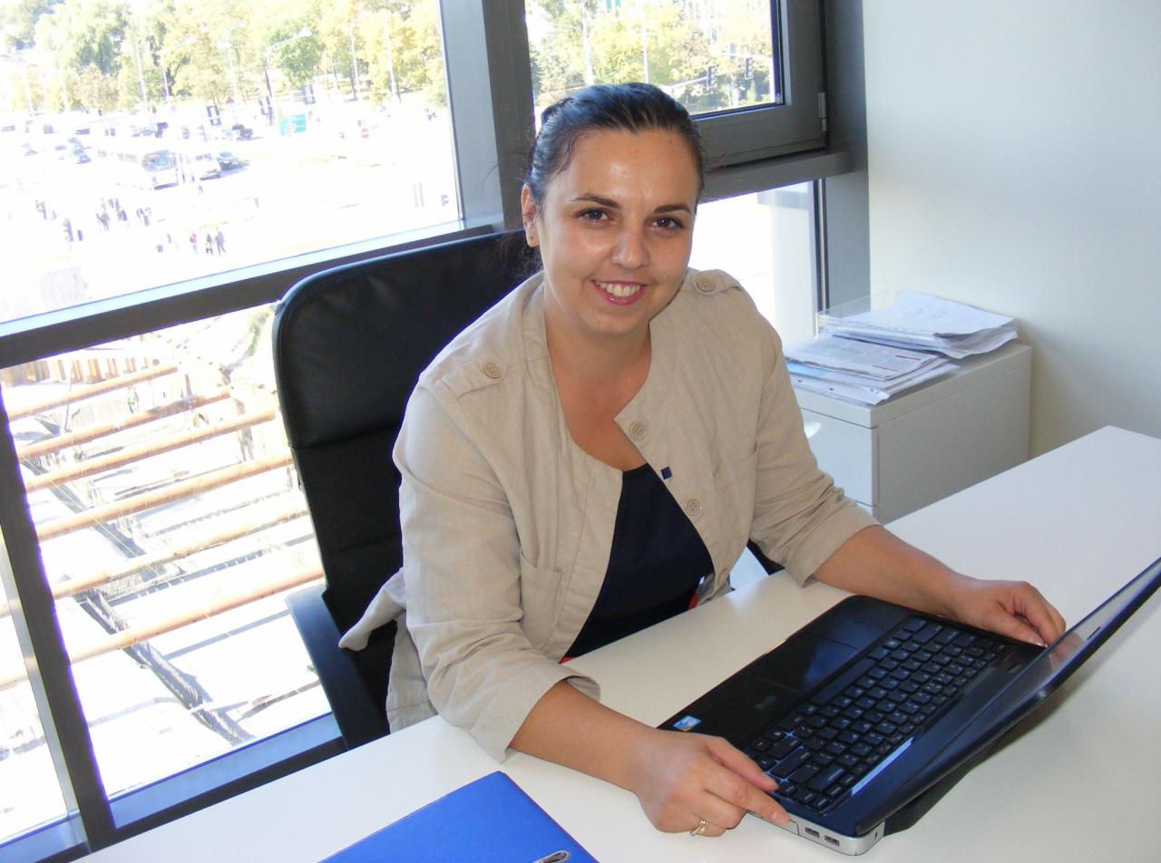 Uśmiechnięta kobieta siedzi za biurkiem z laptopem w jasnym biurze, za nią widok na miasto z okna.