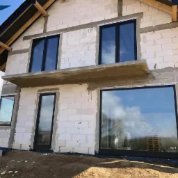 Surowy stan budowy domu jednorodzinnego z zamontowanymi oknami w czarnych ramach, widoczne bloczki z betonu komórkowego i elementy konstrukcji dachu.