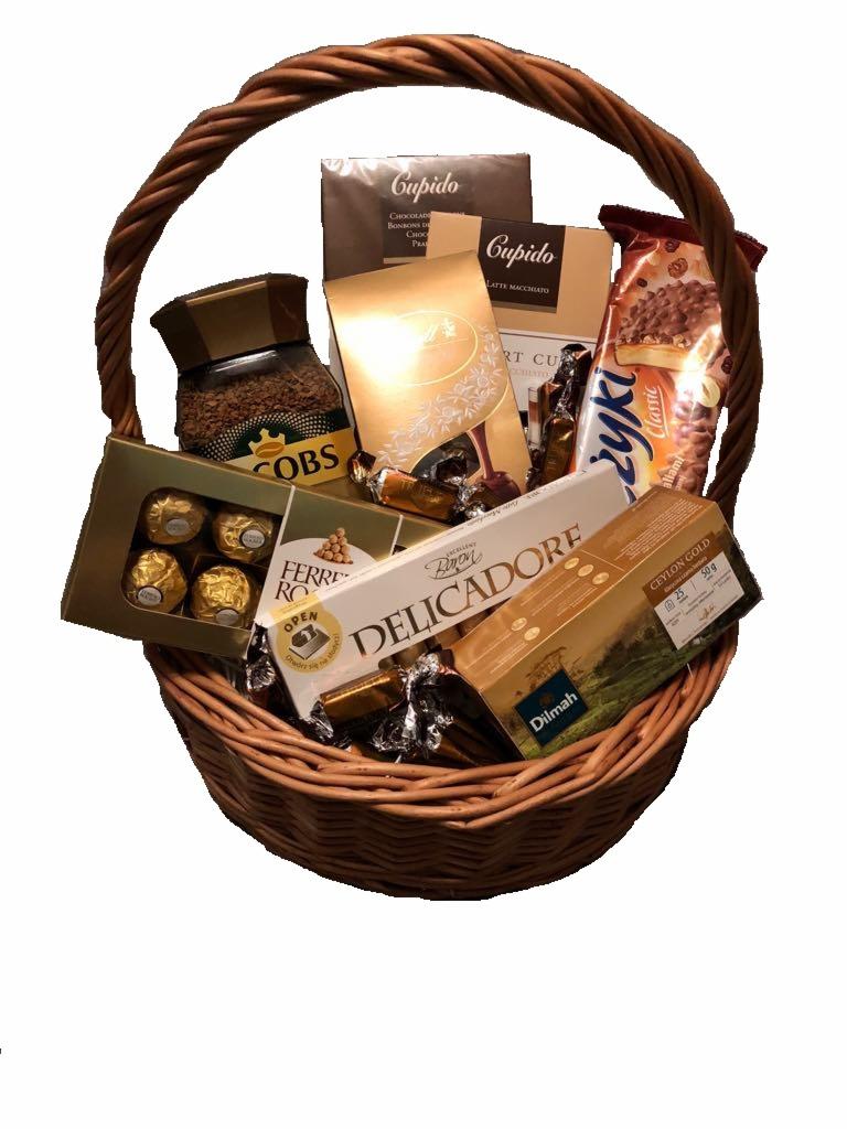 Wiklinowy kosz wypełniony różnorodnymi słodyczami, kawą i herbatą, zapakowanymi w ozdobne pudełka i folie, tworząc elegancki zestaw upominkowy.