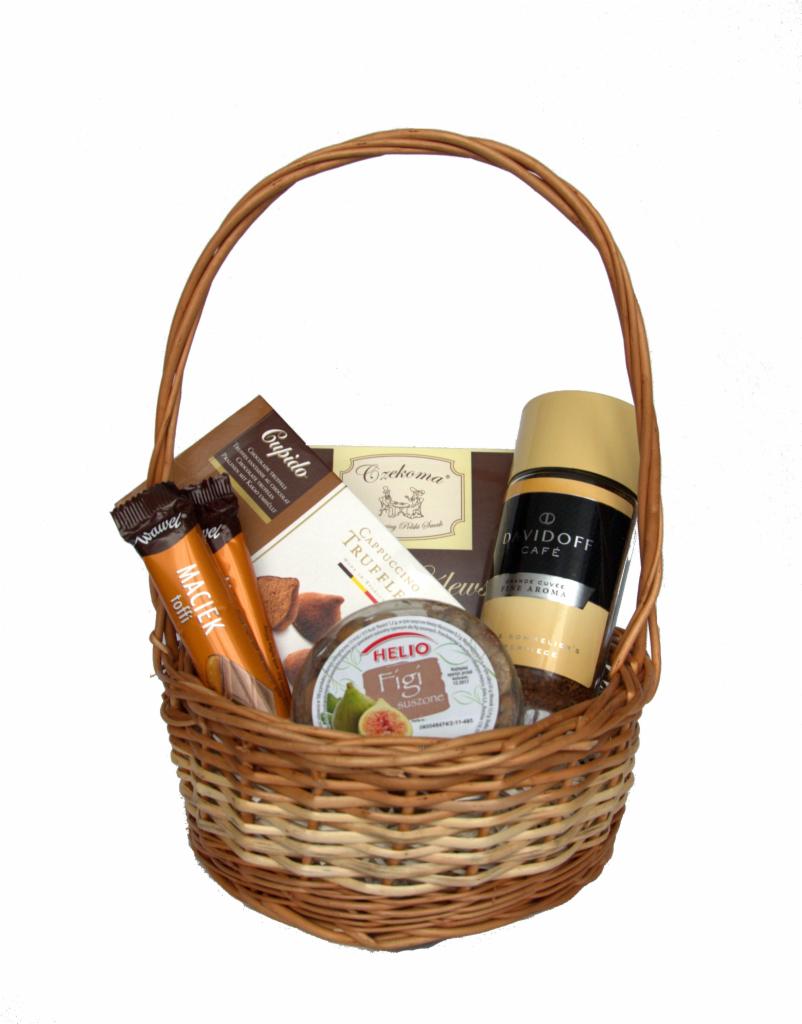 Wiklinowy kosz wypełniony produktami spożywczymi: batoniki Wawel Maciek, czekoladki Cupido, pudełko czekoladek Czekoma, suszone figi Helio i kawa Davidoff, wszystko na białym tle.