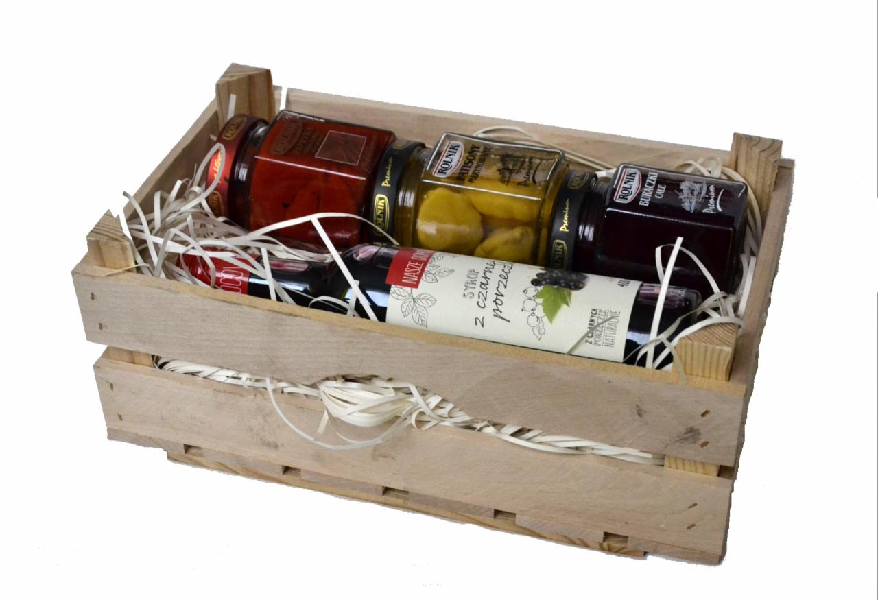 Drewniana skrzynka wypełniona białym wypełniaczem, zawierająca słoiki z przetworami warzywnymi i syropem z czarnej porzeczki marki Rolnik.