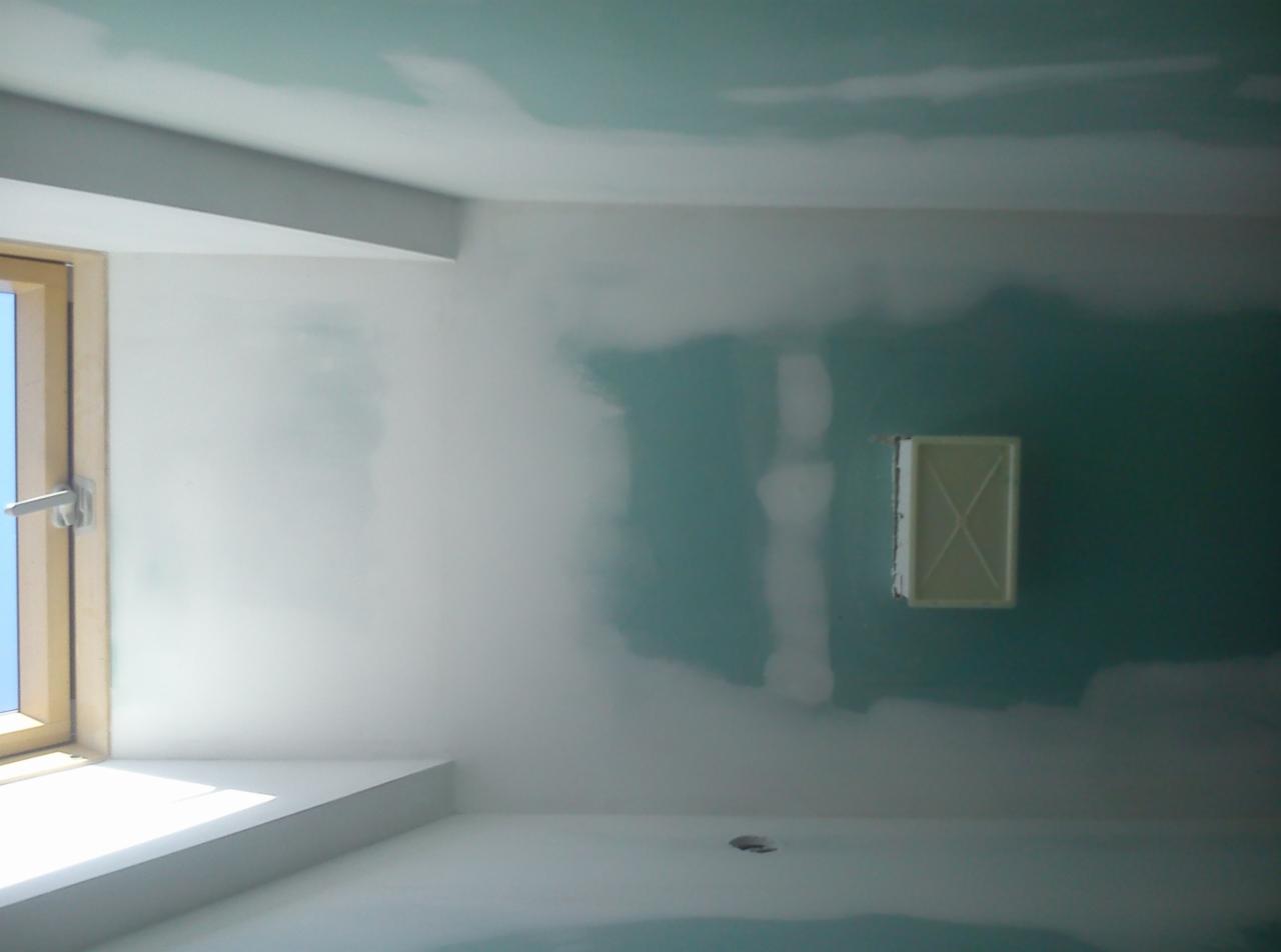 Wnętrze w trakcie remontu: ściany pokryte warstwami gładzi gipsowej, widoczne łączenia płyt kartonowo-gipsowych, okno z drewnianą ramą i kratka wentylacyjna w ścianie.