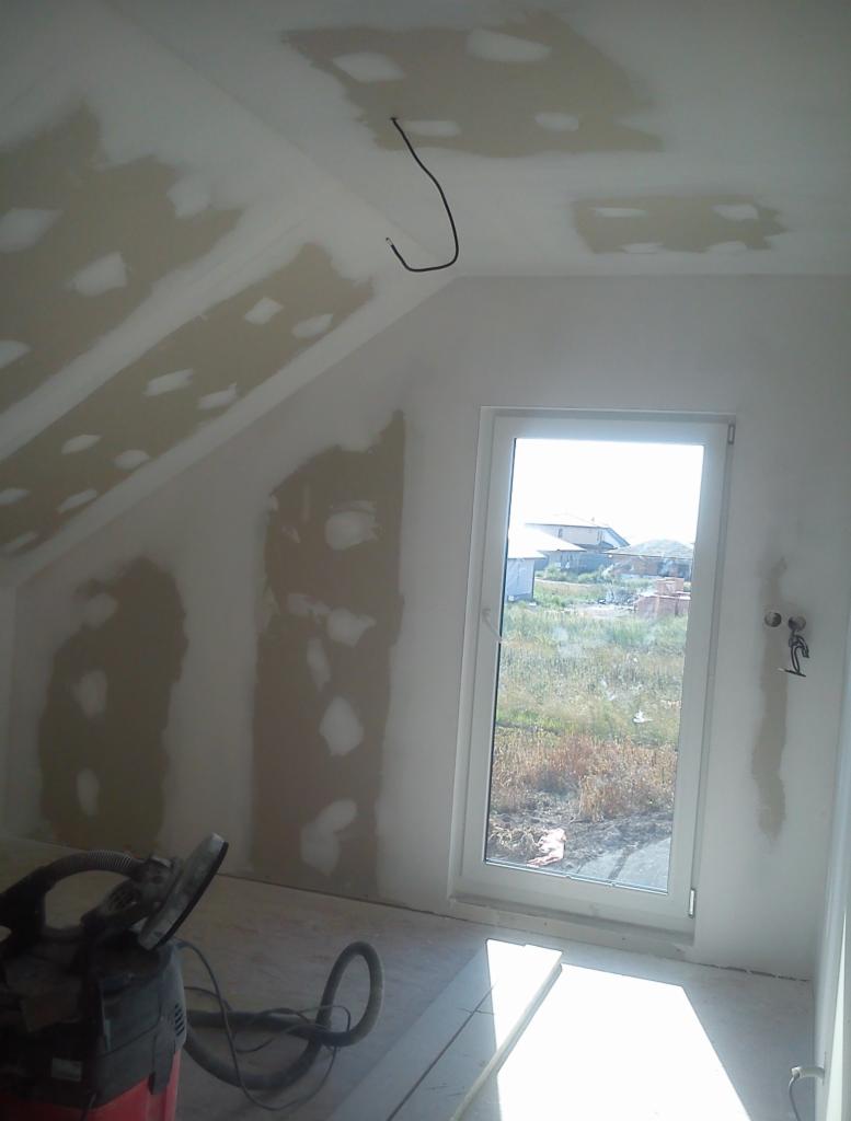 Poddasze w trakcie remontu: ściany i skośny sufit z widocznymi łatami szpachli, kabel zwisający z sufitu, okno z widokiem na zewnątrz, szlifierka do gładzi na podłodze.