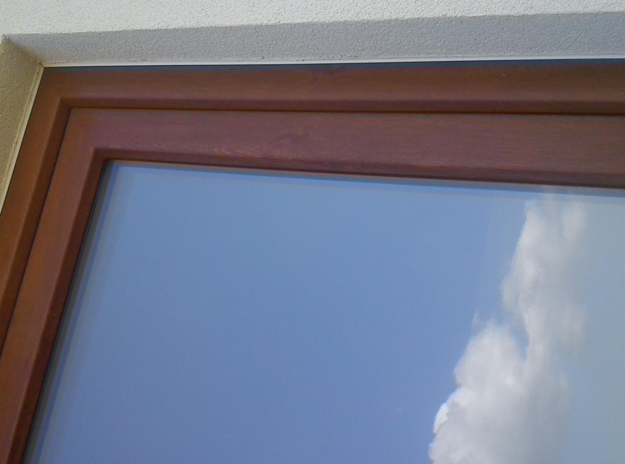 Fragment okna z brązową ramą, widoczna elewacja budynku z jasnym tynkiem oraz odbicie błękitnego nieba z chmurami w szybie.