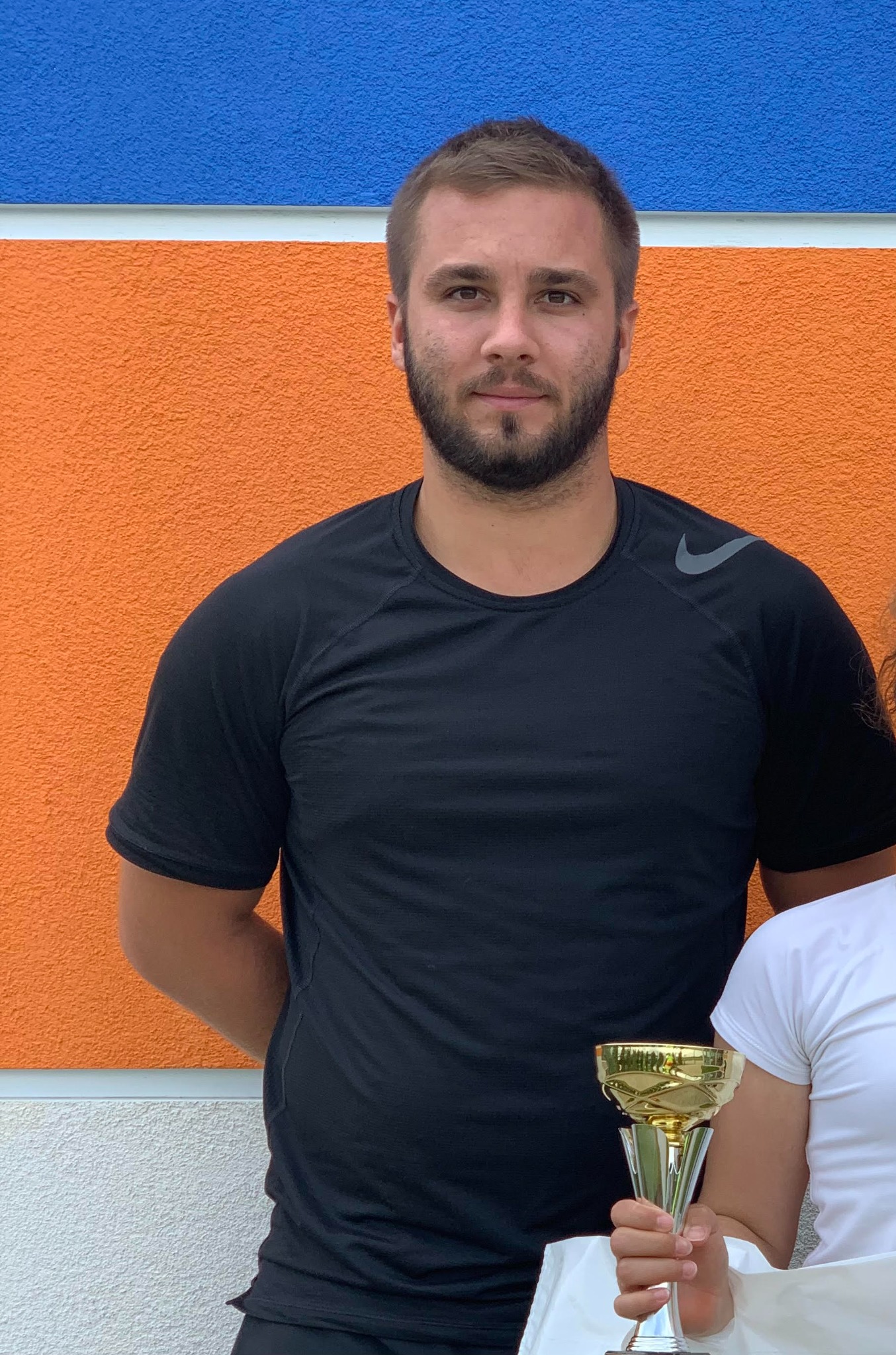 Mężczyzna z brodą w czarnej koszulce z logo Nike, stojący na tle pomalowanej na pomarańczowo-niebiesko ściany, trzymając w ręku złoty puchar.