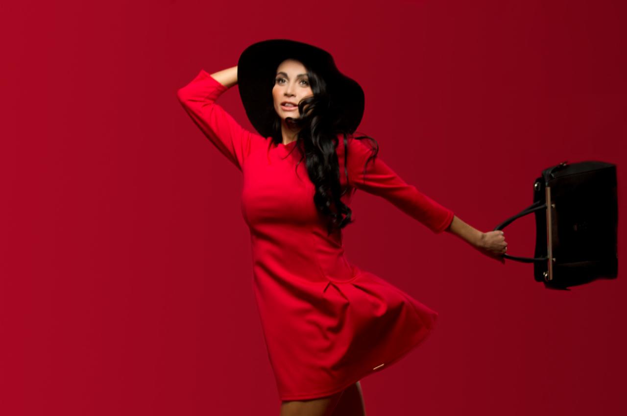 Kobieta w czerwonej sukience i czarnym kapeluszu pozuje z czarną torbą na czerwonym tle.