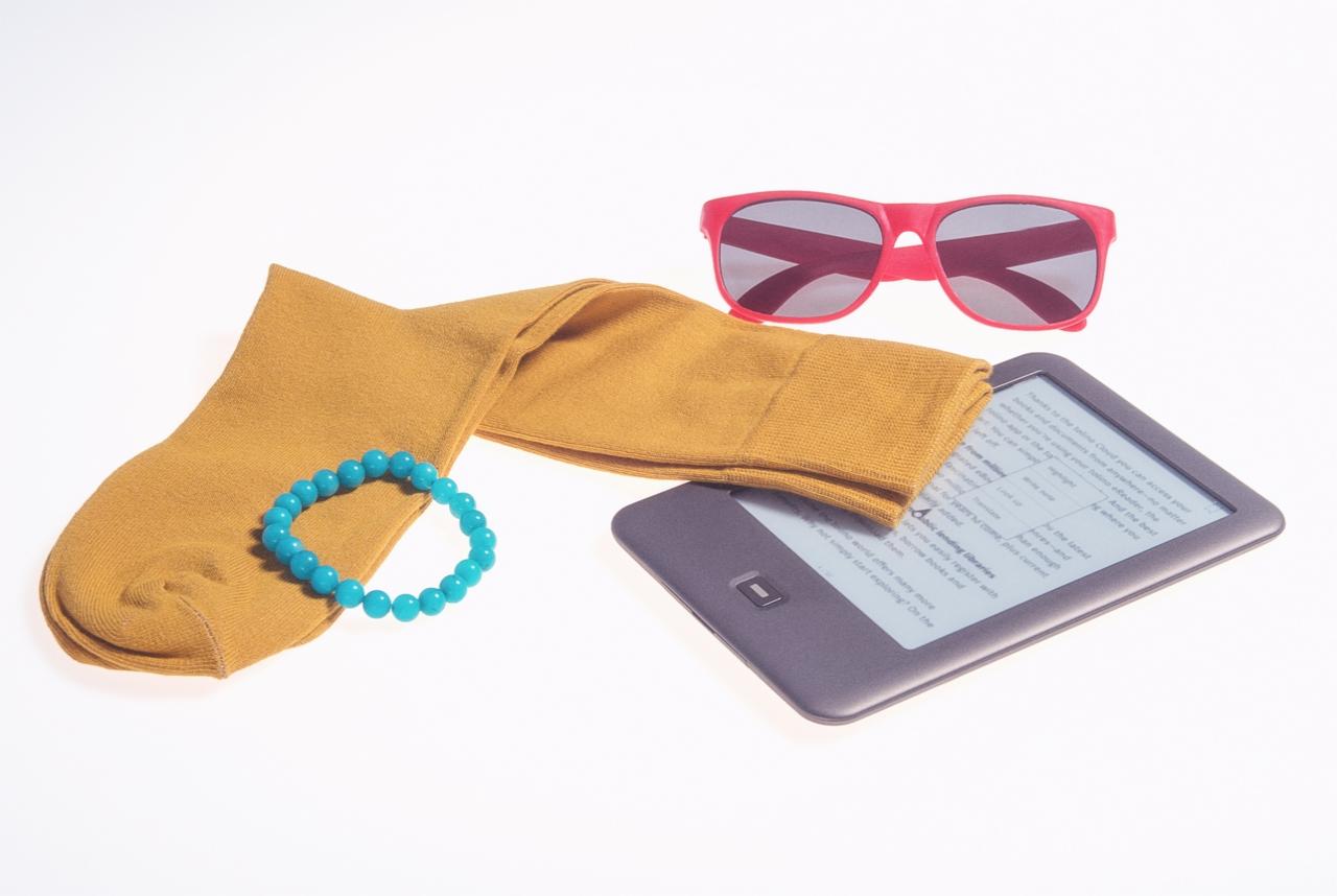 Martwa natura z bransoletką z turkusowych koralików, parą czerwonych okularów przeciwsłonecznych, parą musztardowych skarpet i czytnikiem e-booków na białym tle.
